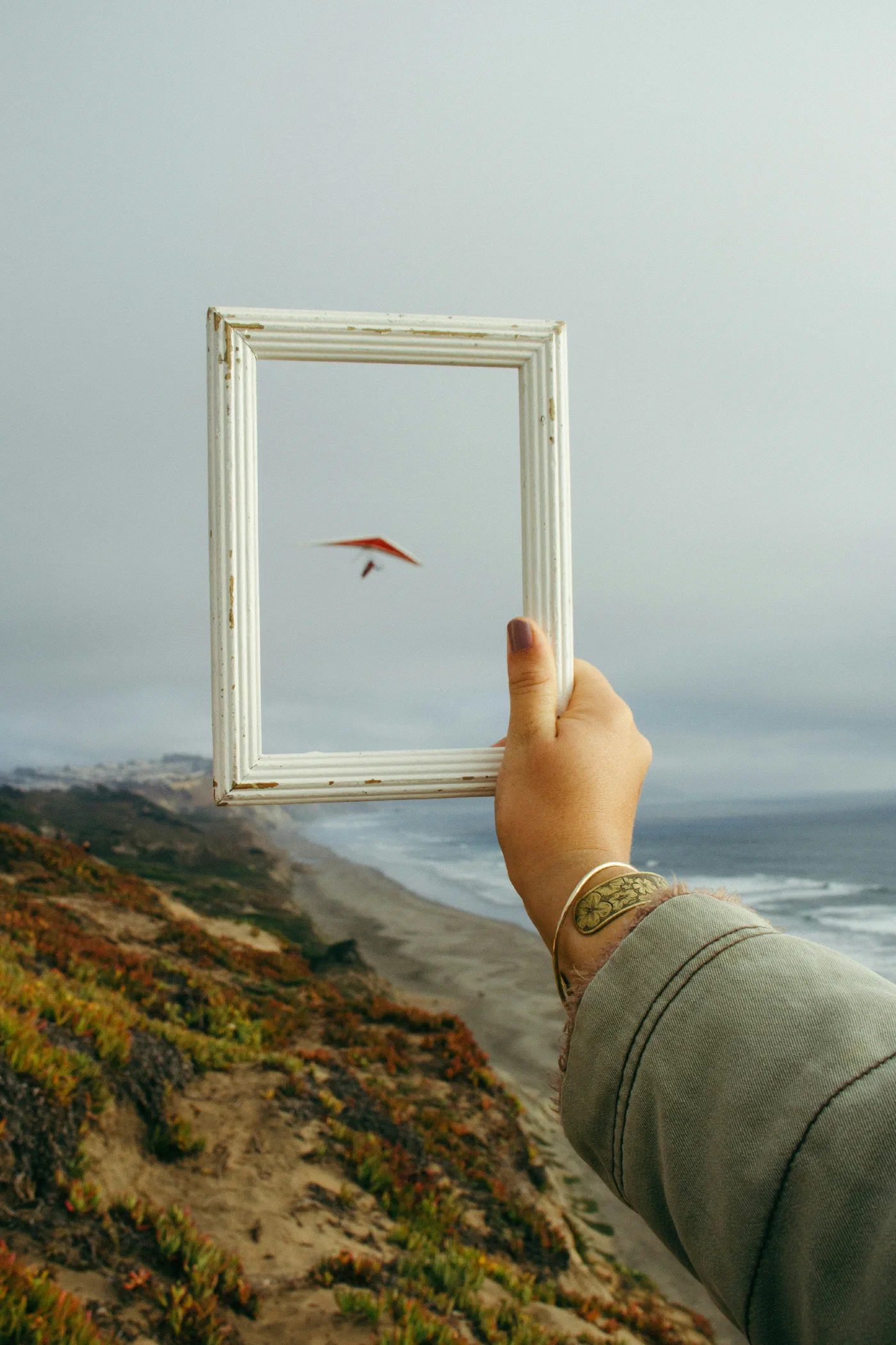 A hand holding an empty picture frame up to a hang glider over a coastal cliff