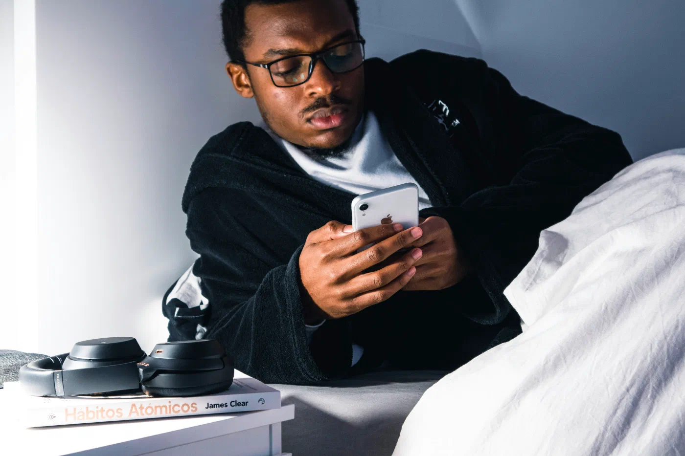 A man in bed scrolling his phone with a copy of Atomic Habits on the nightstand